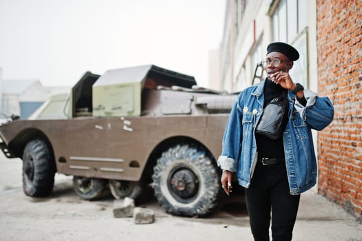 African american man in jeans jacket, beret and eyeglasses, smoking cigar and posed against btr military armored vehicle. photo
