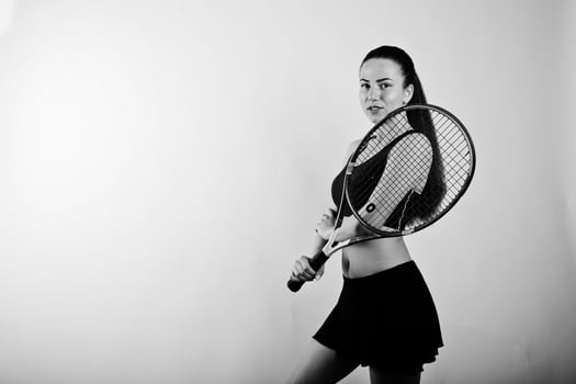 Black and white portrait of beautiful young woman player in sports clothes holding tennis racket while standing against white background. photo