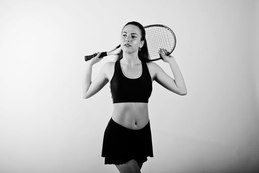 Black and white portrait of beautiful young woman player in sports clothes holding tennis racket while standing against white background. photo