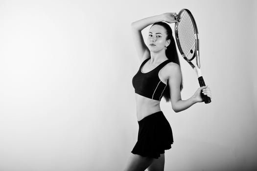 Black and white portrait of beautiful young woman player in sports clothes holding tennis racket while standing against white background. photo