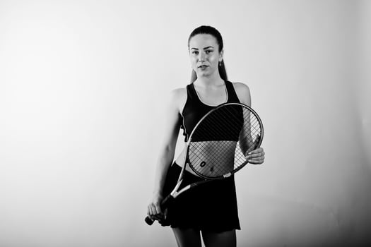 Black and white portrait of beautiful young woman player in sports clothes holding tennis racket while standing against white background. photo