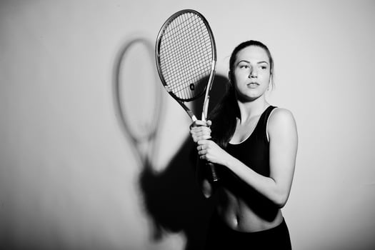 Black and white portrait of beautiful young woman player in sports clothes holding tennis racket while standing against white background. photo