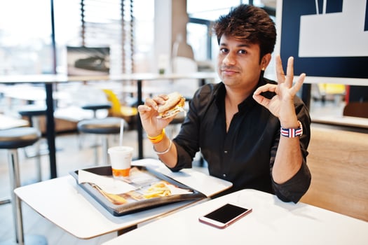 Stylish indian man sitting at fast food cafe and eating hamburger and gesture ok sign. photo