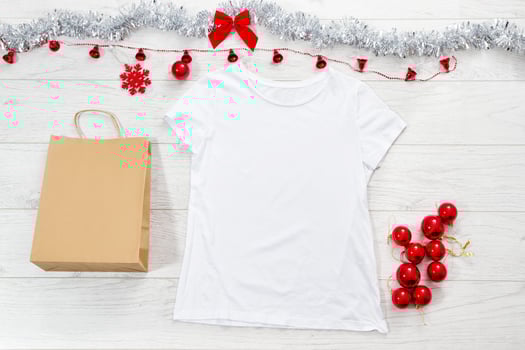 Close up white blank template t shirt with copy space paper bag. Christmas Holiday concept. Top view mockup t-shirt, star, white wooden background. Happy New Year decorations accessories. Xmas outfit photo