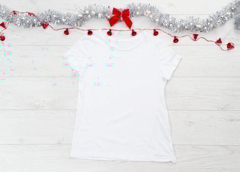 Close up white blank template t shirt with copy space and Christmas Holiday concept. Top view mockup t-shirt and red bow on white wooden background. Happy New Year decorations accessories. Xmas outfit photo