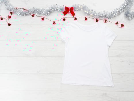 Close up white blank template t shirt with copy space and Christmas Holiday concept. Top view mockup t-shirt and red bow on white wooden background. Happy New Year decorations accessories. Xmas outfit photo