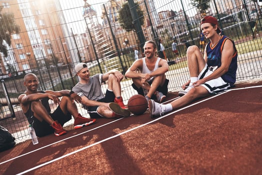 Friends no matter what. Group of young men in sports clothing smiling while sitting on the basketball field outdoors photo