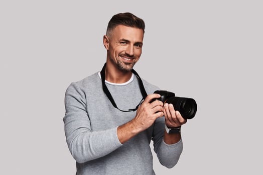 His best hobby. Good looking young man holding digital camera and smiling while standing against grey background photo