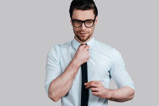 Perfect in his style.  Handsome young man in formalwear looking at camera and adjusting his tie while standing against grey background photo