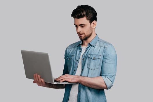 Working on laptop. Handsome young man in blue jeans shirt holding laptop and typing something while standing against grey background photo