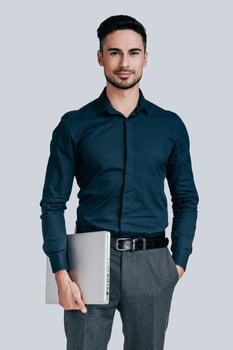 Confident businessman. Good looking young man in shirt carrying laptop and looking at camera with smile while standing against grey background photo