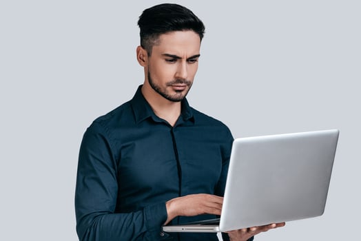 Concentrated at work.  Good looking young man in shirt working on laptop while standing against grey background photo