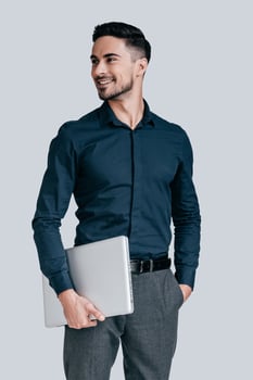 Successful and handsome. Good looking young man in shirt carrying laptop and looking away with smile while standing against grey background photo