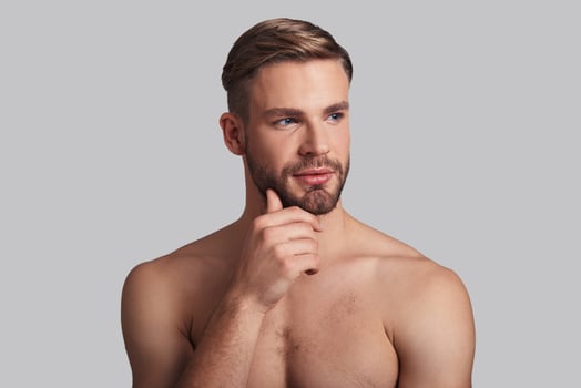 Pleasant dreams. Thoughtful young man looking away and keeping hand on chin while standing against grey background photo
