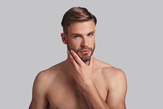 He got the look. Thoughtful young man looking at camera and keeping hand on chin while standing against grey background photo