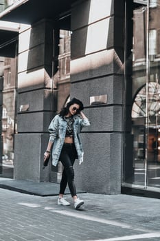Street style. Full length of beautiful young woman in jeans shirt keeping hand in hair while walking down the street photo