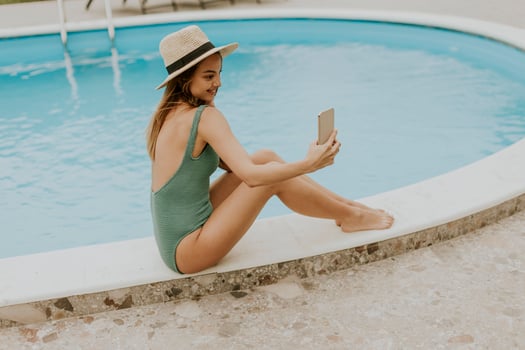 Young woman sitting by the swimming pool and taking selfie photo with  mobile phone in the house backyard