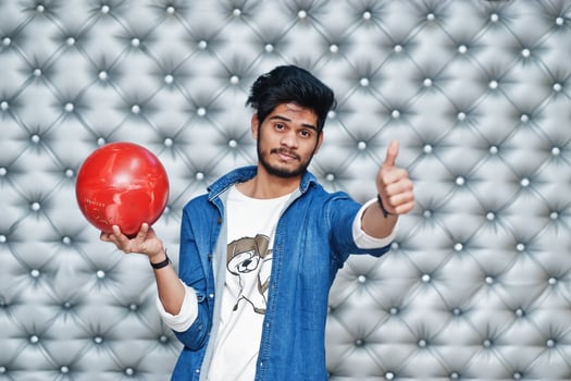 Stylish asian man in jeans shirt standing with bowling ball at hand against silver wall background and show thumb up. photo