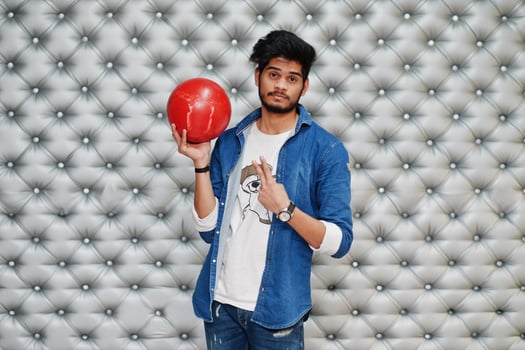 Stylish asian man in jeans shirt standing with bowling ball at hand against silver wall background and show two fingers. photo