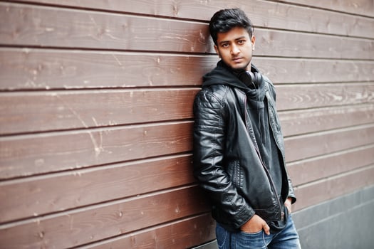 Stylish and casual asian man in black leather jacket, headphones posed against wooden wall. photo