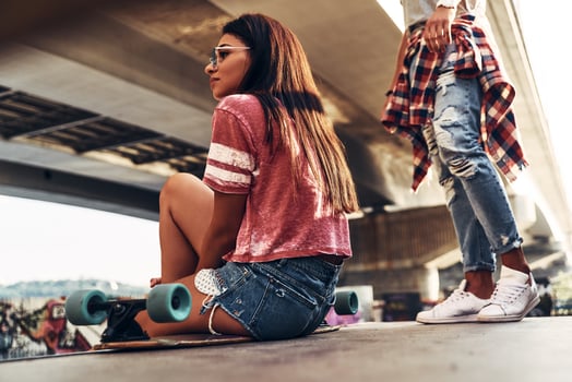 Carefree day. Beautiful young woman sitting on the skateboard and looking away while spending time at the skate park with her friend photo