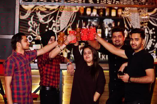 Group of indian friends having fun and rest at night club, drinking cocktails, clinking and cheers near bar counter. photo