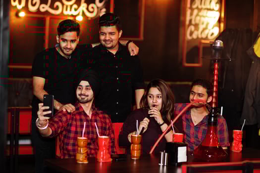 Group of indian friends having fun and rest at night club, drinking cocktails and smoke hookah, looking at mobile phones and making selfie. photo