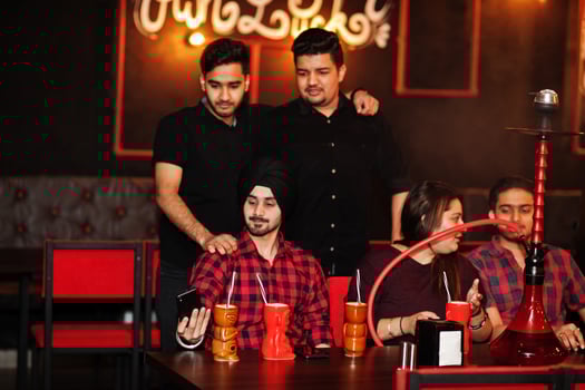 Group of indian friends having fun and rest at night club, drinking cocktails and smoke hookah, looking at mobile phones. photo