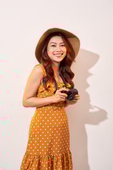 Portrait of cheerful smiling young woman taking photo with inspiration and wearing summer dress. Girl holding retro camera. Model posing on beige background in hat