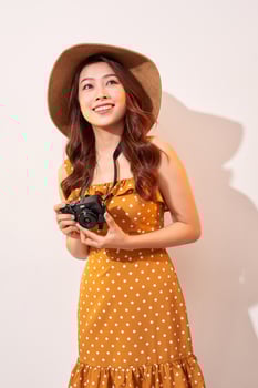 Portrait of cheerful smiling young woman taking photo with inspiration and wearing summer dress. Girl holding retro camera. Model posing on beige background in hat