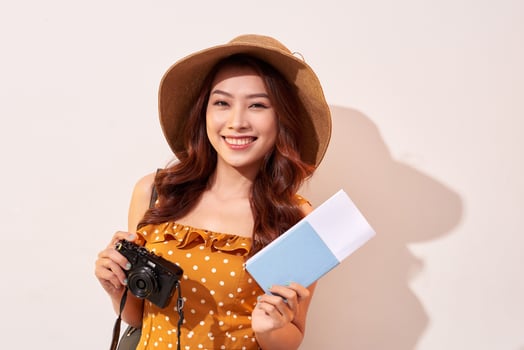 Portrait of a happy young woman in hat holding camera and showing passport while standing isolated over beige background photo