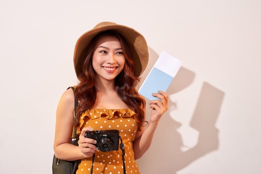 Portrait of a happy young woman in hat holding camera and showing passport while standing isolated over beige background photo