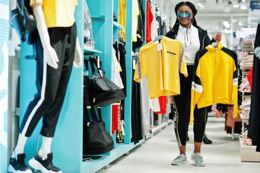 Afican american women in tracksuits and sunglasses shopping at sportswear mall against shelves. She choose yellow t-shirt. Sport store theme. photo
