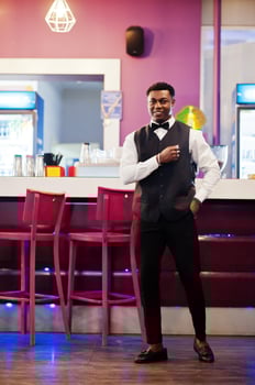 Young handsome african man wearing white shirt, black vest and bow tie posed against bar counter at night club. photo