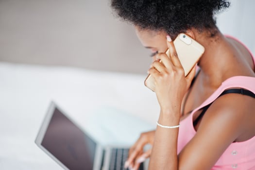 Young african american woman sitting in bed while working on laptop, speaking at mobile phone. photo