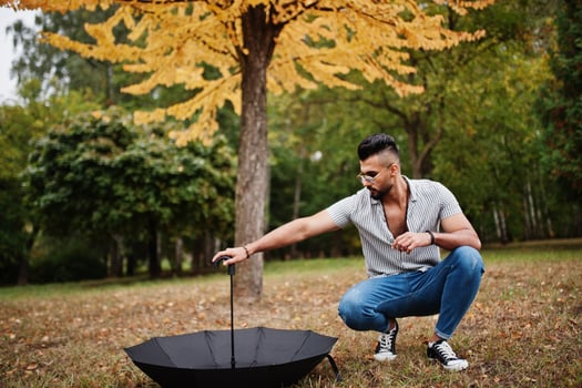 Fashionable tall arab beard man wear on shirt, jeans and sunglasses posed on autumn park with umbrella against yellow leaves tree. photo