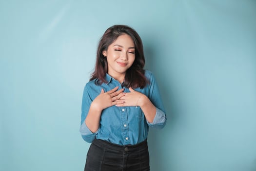 Happy mindful thankful young woman holding hands on chest smiling isolated on blue background feeling no stress, gratitude, mental health balance, peace of mind concept. photo