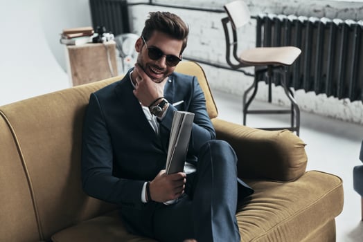 He got candid smile. Handsome young man in full suit keeping hand on chin and smiling while sitting on the sofa photo