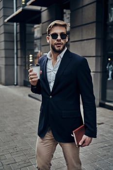 Always in a hurry. Handsome young man in smart casual wear carrying disposable cup while walking through the city street photo