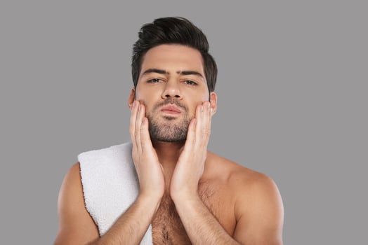 Taking care of his skin. Handsome young man looking at camera and touching his face while standing against grey background photo