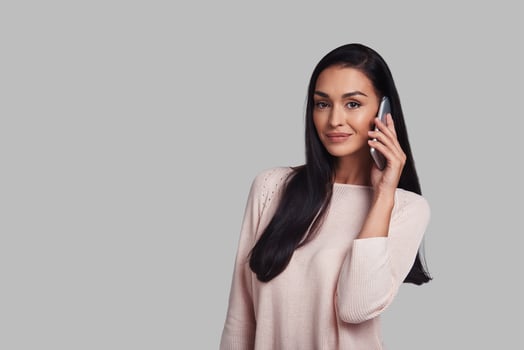 Pleasant talk. Attractive young woman talking on smart phone and smiling while standing against grey background photo