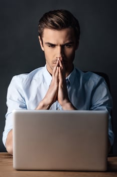 Double-checking his work. Confident handsome young man looking at his laptop and keeping handsclasped while sitting against black background photo