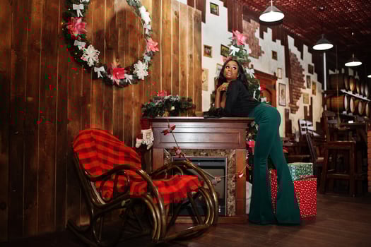 Beauty slim african american model wear on black blouse and green long legs pants posed against christmas mood decorations with wreath, fireplace and rocking chair. photo