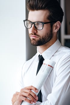Analyzing media news. Pensive young businessman in glasses holding rolled up newspaper and looking away while standing in office photo