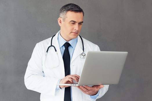 Looking through medical results. Mature male doctor using his laptop while standing against grey background photo