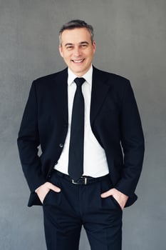 Professional and successful. Mature businessman keeping hands in pockets and looking at camera with smile while standing against grey background photo