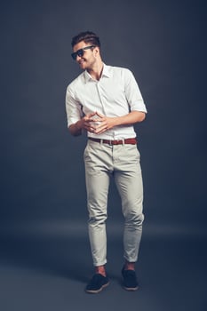 Cheerful and trendy. Full length of confident young handsome man in sunglasses keeping hands clasped and looking away with smile while standing against grey background photo