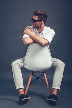 Mr Style. Full length of confident young handsome man in sunglasses looking away while sitting in chair against grey background photo