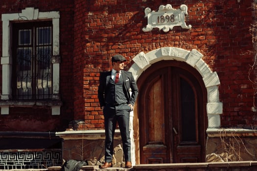 Portrait of retro 1920s english arabian business man wearing dark suit, tie and flat cap standing against old brick house 1898 year. photo