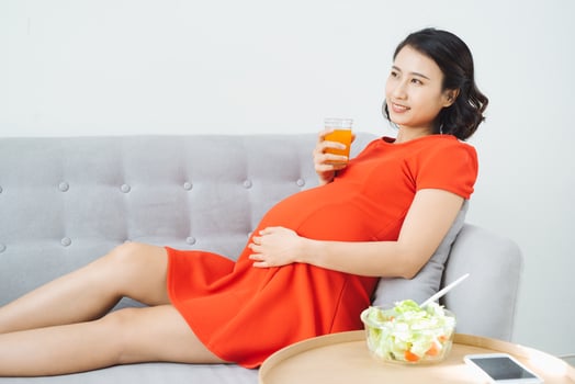 Pretty young Asian pregnant woman eating salad and drinking milk when sitting on sofa. photo